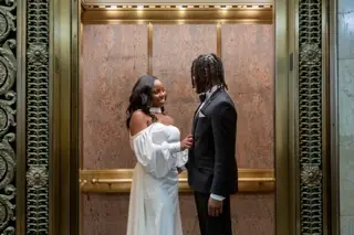 A couple facing each other inside a brass lift, she in a white off the shoulder gown