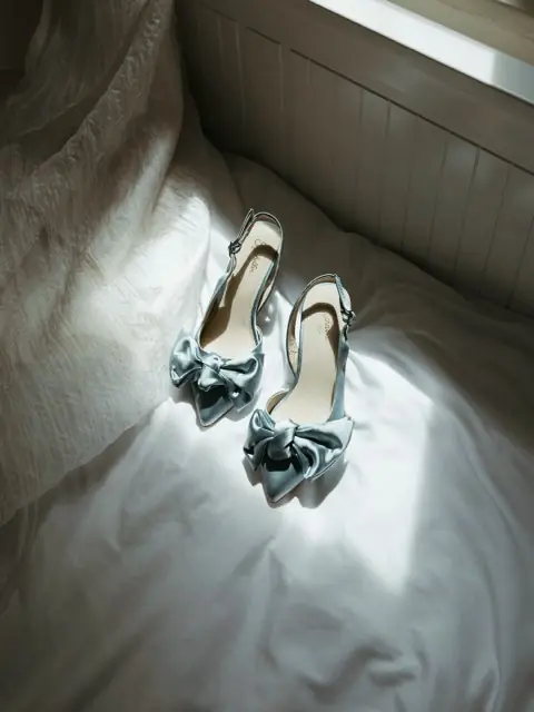 A pair of pale blue satin heels with bows, set on white bedding in window light