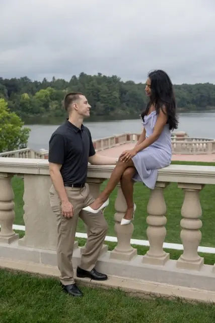 A couple by a lake, she sitting on a stone balustrade and he leaning in towards her