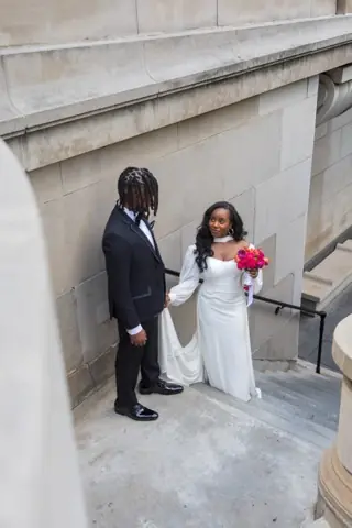 A bride in a white gown holding a hot pink bouquet beside a groom in a black tuxedo on stone steps