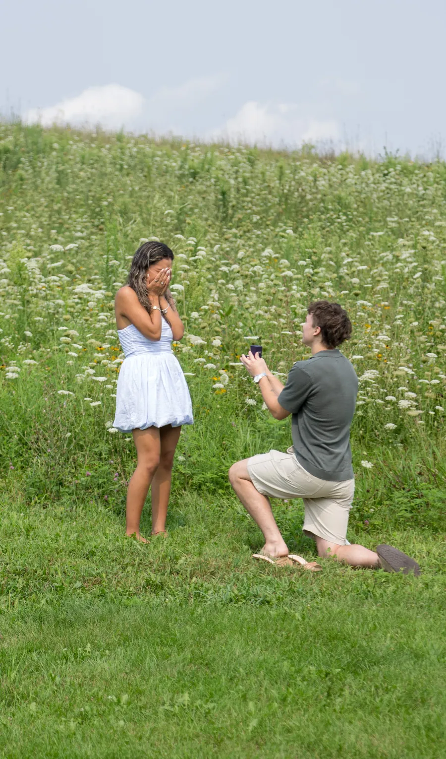 A man on one knee in a wildflower field as she raises both hands to her face
