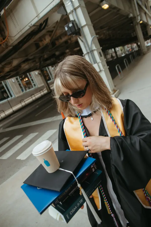 A graduate in sunglasses balancing books, coffee and her cap