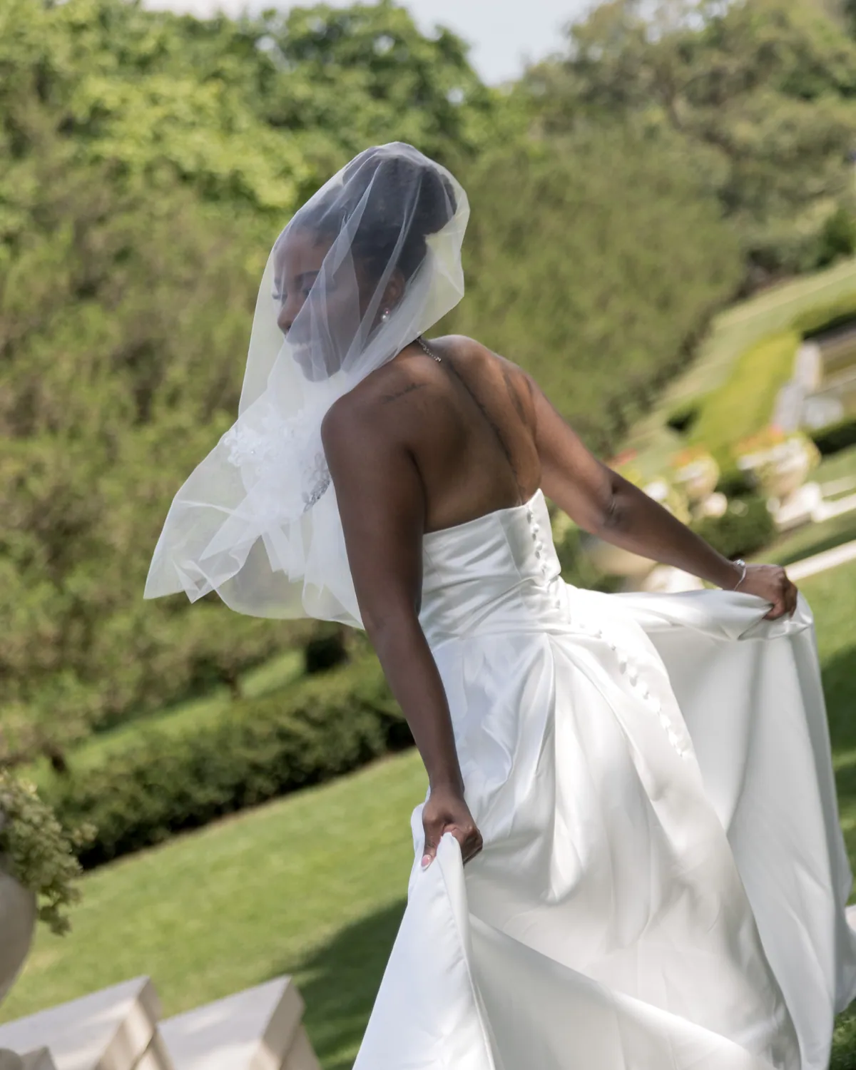 The bride walking away down a clipped box hedge path, veil lifting behind her