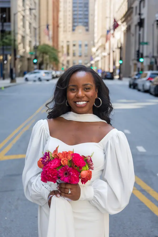 The bride in a long sleeved gown holding a bouquet of hot pink and red blooms, city buildings soft behind her