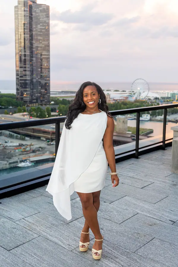 The bride on a rooftop terrace in a short white reception dress, the river and high rises behind her