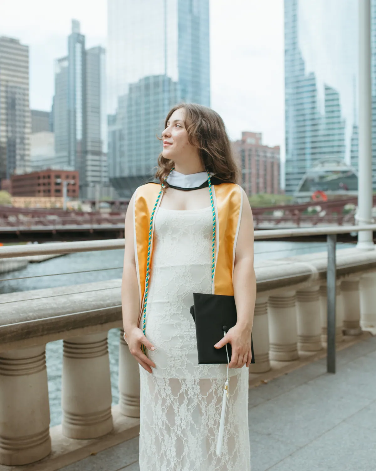 The graduate on the river walk in a lace dress with her gown open over it, the towers of the loop behind her
