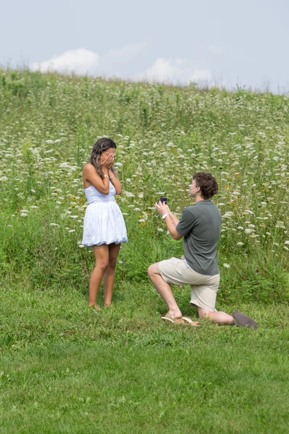 A man on one knee in a tall grass field on a slope while she raises both hands to her mouth